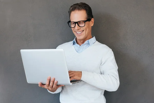 Unterstützung. selbstbewusster, reifer Mann, der vor grauem Hintergrund am Laptop arbeitet und lächelt — Stockfoto