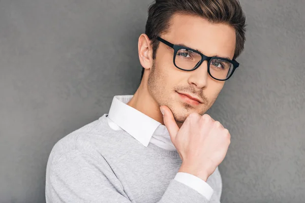Confident and intelligent. Confident young man in glasses holding hand on chin and looking at camera while standing against grey background — Stock Photo, Image