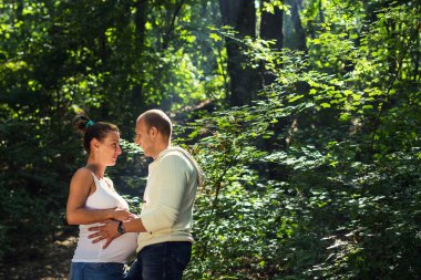 Birbirinin gözlerinin içine bakan ve ellerini hamile kadının karnına koyan çiftin yan fotoğrafı.