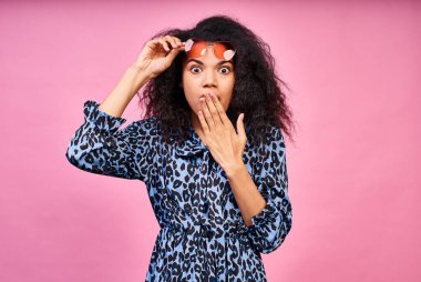 Koyu tenli, afro saçlı, şık kırmızı gözlüklü, mavi elbiseli, gözlüklü ve ağzını avuç içiyle kaplayan genç ve şık moda kızı görünce çok şaşırdı..
