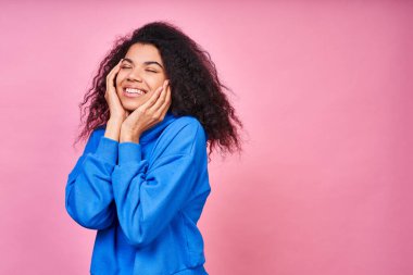 Güzel, genç bir melez, uzun kıvırcık saçlı, mavi bir kapüşonlu giyiyor, rüyasında gözlerini kapatıyor, ellerini yüzünde tutuyor, beyaz bir gülücük. Boşluğu kopyala.