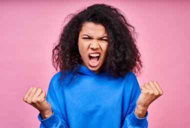 Yakın plan, gür kıvırcık siyah saçlı, mavi ceketli, ellerini yumruklarına sıkıştırmış, yüksek sesle bağıran, düz bakışlı, pembe bir arka planda poz veren genç bir kız..