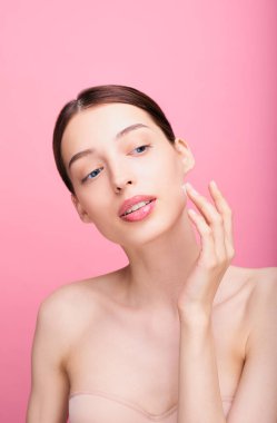 Güzel surat. Doğal makyajı ve sağlıklı deri portresi olan bir kadın. Güzel kız model pembe arka planda parlayan hidratlı cilde dokunuyor. Cilt bakımı kavramı.