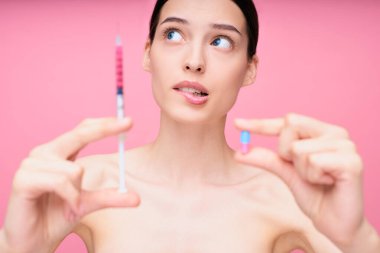 Sevimli genç bir kız elinde şırınga ve elinde hapla bir seçim yapar, dudağını ısırır ve başını çevirir. Kozmetik ve enjeksiyon cerrahisi, ilaçlar, ilaç konsepti.