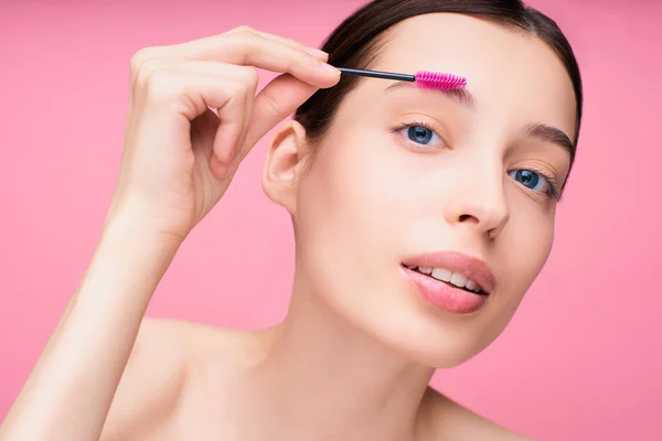 Eyebrow makeup. Woman brushing eyebrows with brush close-up. Model girl with beautiful blue eyes and dark hair shaping her eyebrows with eyebrow gel on a pink background.