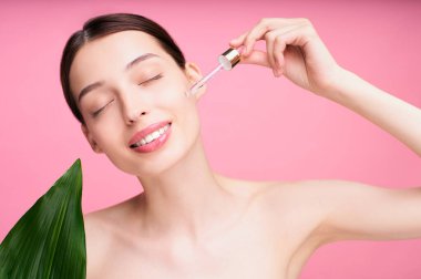 Çekici bir genç kız pipetle yüzüne hyaluronik serum uyguluyor. Yeşil tropikal yapraklı, pembe arka planda makyajsız çekici bir kadının fotoğrafı. Güzellik konsepti.