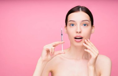Şaşırmış, korkmuş, çekici beyaz bir kız model yüzünde botokos olan bir şırınga tutuyor. Yetişkin bir kıza pembe arka planda kozmetik iğne yapılıyor. Plastik Cerrahi Kliniği.