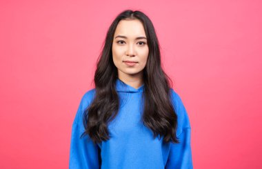 Güzel bir Asyalı kadın, tatlı bir gülümsemesi, pembe yanakları, mavi bir kapüşonlu giyiyor, samimi duygularını ifade ediyor, koyu renk saçları var, pembe arka planda soyutlanmış. Mutluluk kavramı.