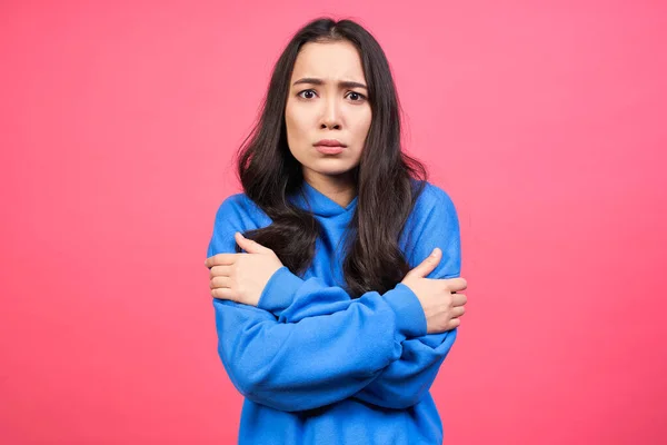 Sad lonely Asian woman with black hair, hugging herself by the shoulders, bored, looks sad and tired, wears a blue sweatshirt with a hood, stands against a pink background. People and sadness.