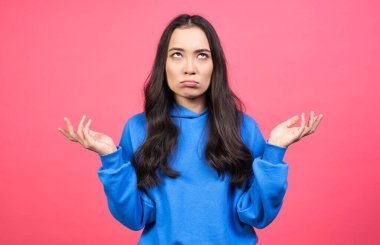 Kafası karışmış genç Asyalı kadın avuçlarını yana doğru açıyor, ne yapacağını bilmiyor, günlük mavi kapüşonlu giyinmiş, kameraya üzgün bakıyor, sorunlu bir durumda nasıl davranacağını düşünüyor..