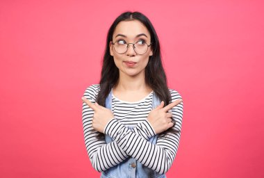 İki ürün arasında seçim yapan Asyalı bir kadın. Kollarını çapraz yapıp karşı tarafı işaret ediyor. Pembe arka planda izole edilmiş günlük kıyafetler giyiyor. Boşluğu kopyala.