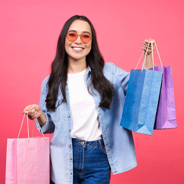 Joyful cheerful beautiful Asian girl in a white T-shirt and denim shirt, holding multi-colored bags, packages, after a successful shopping, isolated on a pink background.