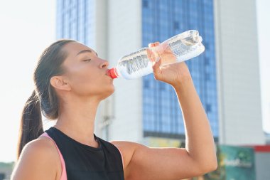 Genç, taze, güzel, narin bir kız, uzun siyah saçlı, siyah bir spor gömleği giyiyor, spor yaptıktan sonra bir şişeden su içmeyi seviyor, şehrin arka planında.