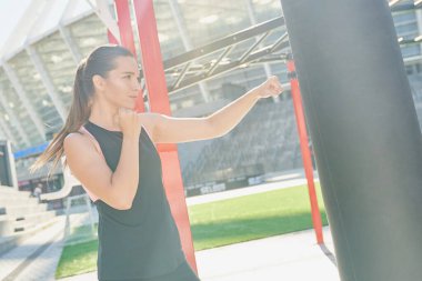 Spor, modern giysiler içinde, açık hava spor sahasında kum torbası tekmeleyen güçlü, kendine güvenen genç bir sporcu. Güçlü ve bağımsız bir kadın, yeni nesil bir kadın, feminizm, eşitlik..