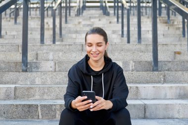 Çekici, sevimli, modern, siyah spor giyimli, uzun siyah saçlı, at kuyruklu, açık havada merdivenlerde oturuyor ve akıllı bir telefona bakıyor. Spor, spor ve teknoloji..