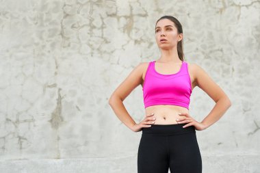 Pembe bluzlu, koyu renk uzun saçlı, açık havada idmandan sonra dinlenen genç bir kadın, elleri belinde gri bir duvarın yanında duruyor. Spor onun yaşam tarzı. Lider kadın..