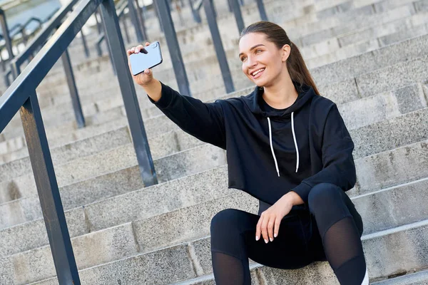 Gülümseyen, neşeli, modern kız atlet, fitness bayan, siyah spor giyim, gri basamaklarda oturuyor ve atletik performansıyla gurur duyan bir selfie çekiyor..