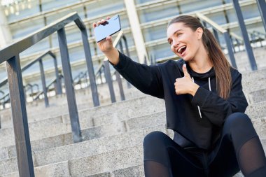 Gülümseyen, neşeli, modern bayan sporcu, spor giyimli bayan sporcu, gri basamaklarda oturuyor ve selfie çekiyor ve işaret gibi görünüyor. Sağlıklı bir yaşam tarzı..