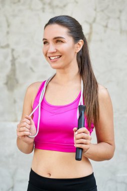 Gülümseyen, mutlu, cana yakın, pembe bir bluz giyen boynuna ip geçirip mutlu bir şekilde uzaklara bakan bir kızın dikey çekimi. Kadın fitness eğitmeni, kadınlar için spor.