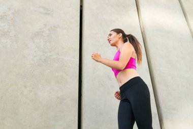 Alt manzara, spor giyimli genç bir kadının gri arka planda kardiyo antrenmanı yaparken görüntüsü. Spor yapan kadınlar yaz sabahları dışarıda egzersiz yaparlar. Sola doğru koşar..