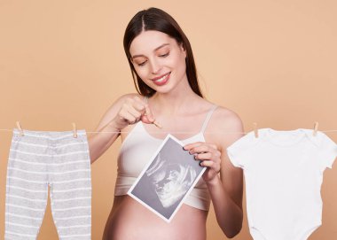 Çekici, nazik, romantik, beyaz bluzlu, uzun siyah saçlı, beyaz saçlı beyaz bir kız, doğuma hazırlanıyor, çocuk kıyafetleri topluyor, seviniyor ve ileriye bakıyor..