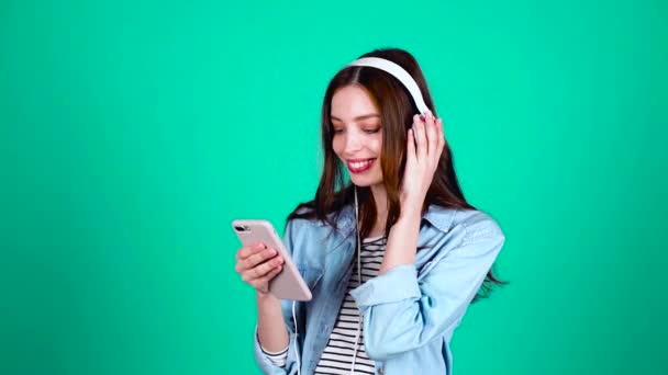 縞模様のセーターとデニムのシャツに身を包んだ10代の可愛い少女は ヘッドフォンで好きな音楽を聴き スマートフォンを手に持っている 人と道具の概念 — ストック動画
