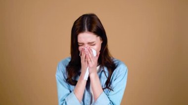 Ağlayan, uzun siyah saçlı, mavi gözlü, modaya uygun kıyafetlerle genç bir kız, göz yaşlarını kağıt peçeteyle siliyor, üzgün, kahverengi bej bir arka planda poz veriyor..