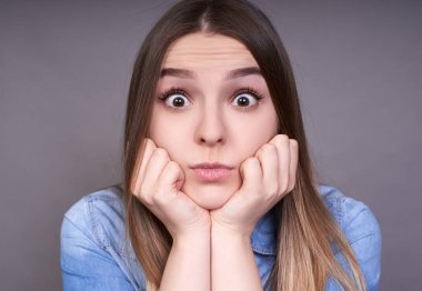 Yakın plan, kahverengi gözlü, uzun saçlı, ellerini çenesinin altında tutan ve gri bir arka planda poz veren genç beyaz bir kız..