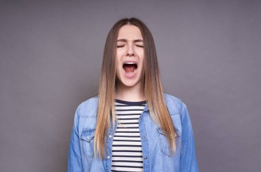 Gözleri kapalı, uzun saçlı, çizgili kazak ve kot gömlekli, gri bir arka planda tek başına duran heyecanlı, üzgün, çığlık atan genç bir kadının görüntüsü..
