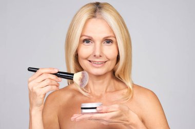 Makyaj yapmak üzere olan, elinde pudra ve makyaj fırçası olan yetişkin, beyaz, çıplak bir kadının fotoğrafını çek. Makyaj fırçasıyla allık süren olgun bir kadın portresi..