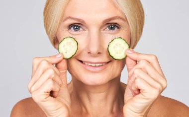 Yakın plan, sarışın ve mavi gözlü orta yaşlı güzel bir kadın yüzüne salatalık sürüp estetik yenileme işlemleri yapıyor. Organik doğal kozmetik konsepti.