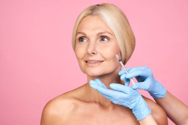 Profesyonel klinikte kırışıklıkları olan, gülümseyen, çekici, yaşlı bir kadın. Elleri eldivenli, pembe arka planda izole edilmiş yüz hatları yapan..
