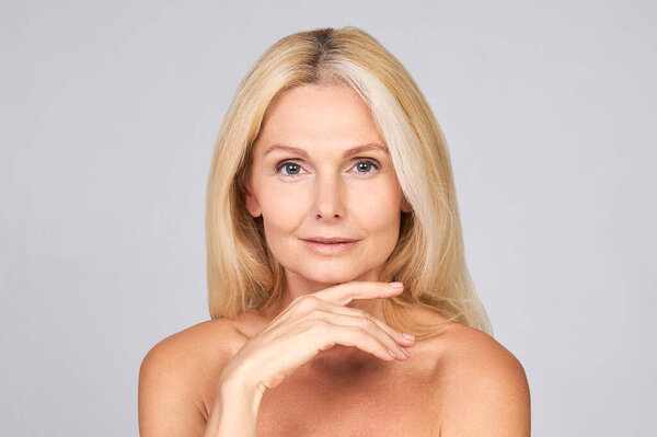 Cute beautiful attractive elderly lady, naked shoulders, perfect face skin, holds a palm under the chin, posing in the studio on a light background. Mature beauty concept, mature skin care.