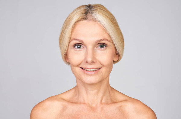 A cute smiling mature blonde with beautiful well-groomed skin, looking straight ahead, posing on a light gray background. The concept of beauty, self-care, mature anti-aging skin care.