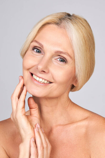 Vertical shot of smiling natural cute pretty mature Caucasian lady with blond tied hair looking at the camera and touching her cheek. Mature skin care concept for mature skin, cosmetology for mature.