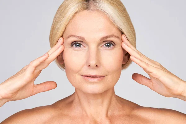 Gri pastel arka planda izole edilmiş, temizleyici ipek tene dokunan sevimli, yaşlı bir kadının yakın plan portresi. Olgun cilt bakımı konsepti, olgun cilt kozmetolojisi.