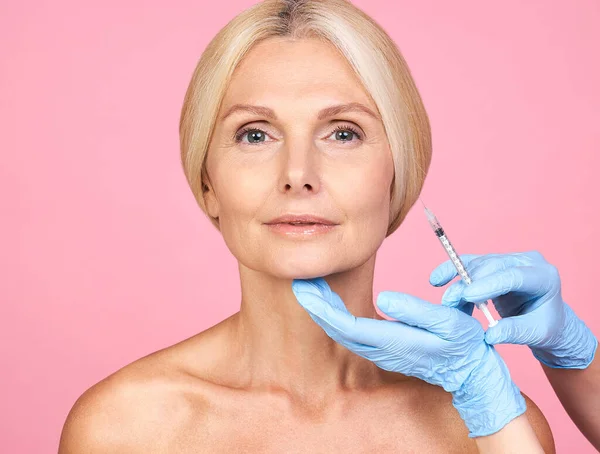 Profesyonel klinikte kırışıklıkları olan çekici bir kadın. Elleri eldivenli, pembe arka planda izole edilmiş yüz hatları yapıyor..