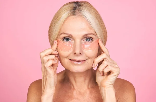 微笑着的中年妇女把粉红的斑斑放在她的眼睛 看着相机 干性皮肤护理美容疗法概念 抗老化 抗皱肌肤更新 — 图库照片