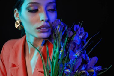 Yakın plan, makyajlı, bir buket çiçekli, parlak bir genç kız siyah arka planda, renkli UV filtreli poz vermekten hoşlanıyor. Özel tasarım, tarz, moda, cazibe.