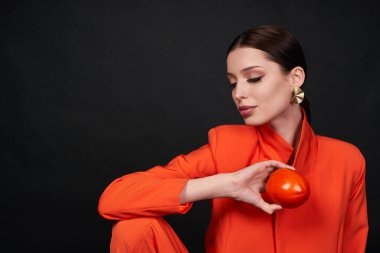 Stüdyoda elinde egzotik bir meyve tutarak şık turuncu bir takım elbiseyle poz veren nazik, genç bir kadın manken. Siyah arkaplandaki fotoğraf. Boşluğu kopyala.