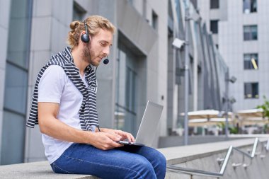 Çekici genç bir adam, serbest çalışan, kot pantolon ve beyaz tişörtlü, uzaktan çalışıyor, iş arkadaşlarıyla sokakta, şehirde bir video konferansı düzenliyor..