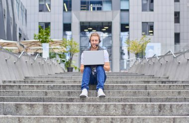 Şehir yaşam tarzı genç adam bir binanın basamaklarında oturuyor dizüstü bilgisayarla online bir klavye yazıyor. Modern yaşam tarzı, iletişim, iş, çevrimiçi öğrenme, serbest çalışma kavramı.