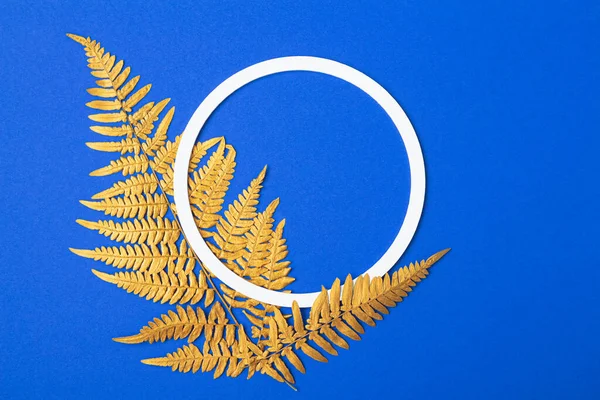 Minimalistic natural composition. White round frame and golden fern leaves on blue background. Copy space.