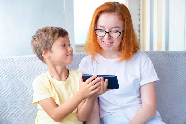 Mother and son sitting on sofa playing on smartphone and smiling, having fun. Modern technologies and Internet in children\'s education and leisure.