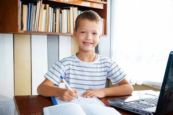 Distance learning online education. Schoolboy boy looks at camera and smiles, studies at home with laptop and does school homework. Training books and notebooks on table