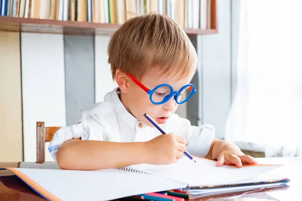 Little boy in funny round glasses is sitting at table and drawing with colored pencils. Concept of early development and preschool education.