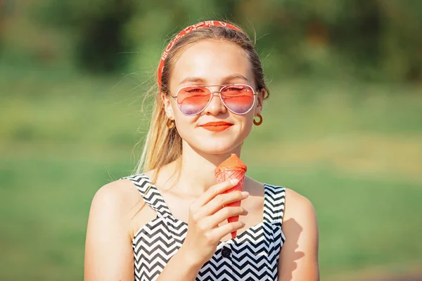 Young blonde hipster girl eating delicious pink ice cream in summer hot weather in sunglasses have fun and good mood and smiling
