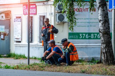 Yol yapımcılarından oluşan bir ekip, bir yaz günü şehir merkezinde kaldırımın kenarında, taş bir binanın yanında dinleniyor.