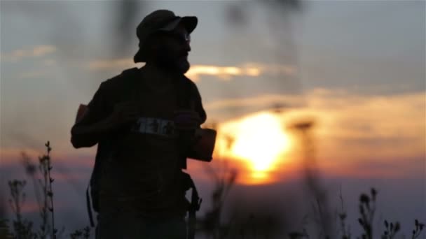 En turist med en ryggsäck och en kamera vid solnedgången i fältet. Skäggig man resenär gör selfie vid solnedgången. — Stockvideo