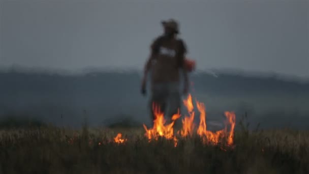 Vousatý Turistický batoh nedaleko hořícího pole. Muž v klobouku a cestovatel s batohem na pozadí plamenů. — Stock video